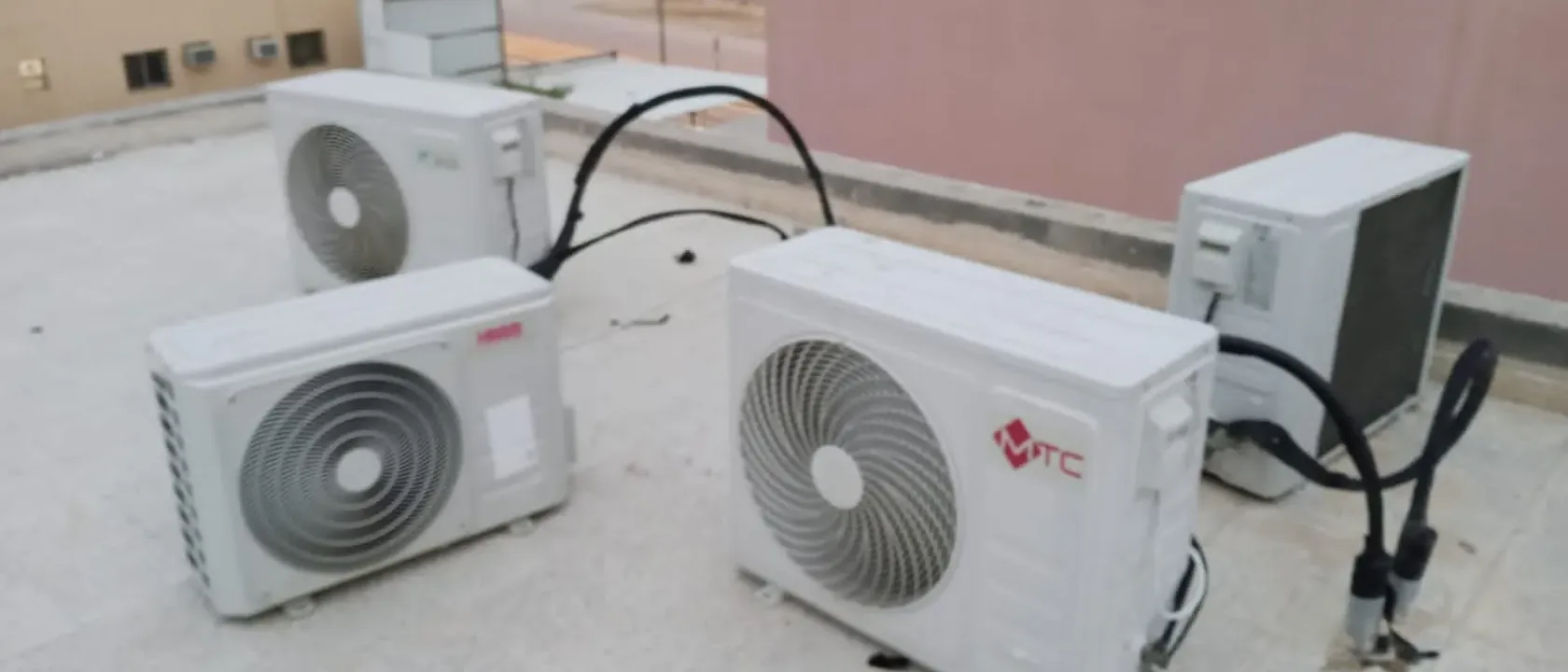 صيانة المكيفات في البيت تشمل ثلاث وحدات تكييف خارجية على سطح منزل بجدران وردية